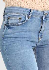 Ljust blå denimjeans med en lätt distressed yta, med en liten frammyntficka, guldsömmar och metalldekorationer.