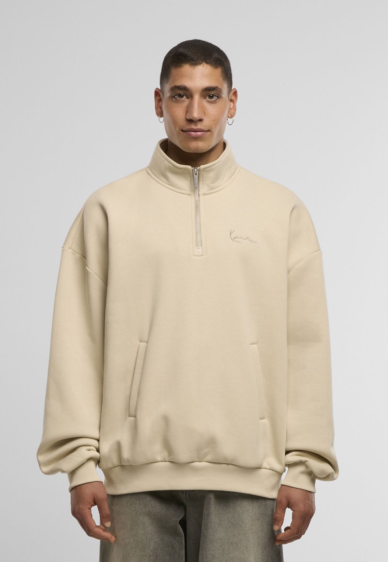 Sweat-shirt beige à fermeture éclair avec un col montant, une coupe oversize et des poches latérales. Comprend un logo brodé subtil sur la poitrine et des poignets côtelés.