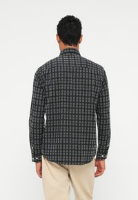 Man die met zijn rug naar de camera staat, gekleed in een zwart-wit geblokte longsleeve shirt en lichtbeige broek, tegen een simpele witte achtergrond.