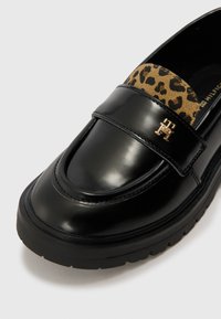 Svart lackläder loafer med ett band i leopardmönstrad mocka och en guldsymbol. Har en tjock gummisula för ökad komfort.