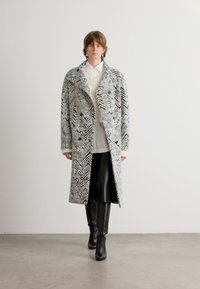 Abrigo largo con patrón de houndstooth en blanco y negro, hecho de una tela texturizada. El modelo lleva una camisa blanca y pantalones negros, con botas hasta la rodilla.