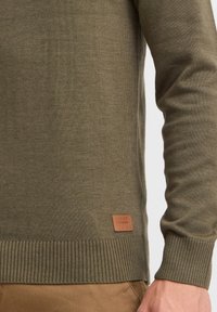 Pull vert olive avec un ourlet et des poignets côtelés, fabriqué en matériau tricoté doux. Une étiquette en cuir marron avec une marque est visible en bas.
