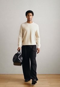 Bolsa de lona negra con textura sostenida en una mano; el modelo lleva un suéter crema y pantalones negros sueltos, de pie sobre un suelo de madera.