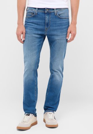 Mann trägt hellblaue Slim-Fit-Jeans und weiße Sneakers, steht vor einem einfarbigen Hintergrund.