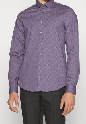 Mężczyzna ubrany w dopasowaną, fioletową, wzorzystą koszulę na guziki z długim rękawem oraz czarne spodnie, pokazany od szyi do bioder.