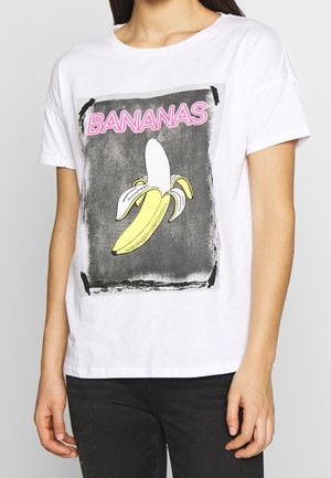 Osoba ubrana w białą koszulkę z nadrukiem obranego żółtego banana oraz słowem "BANANAS" różowym kolorem nad nim na szarym tle.