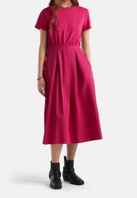 Nainen, joka pukeutuu kirkkaan vaaleanpunaiseen midi-mekkoon, jossa on lyhyet hihat, ja mustiin nilkkasaappaisiin, seisoo yksi käsi taskussa valkoista taustaa vasten.