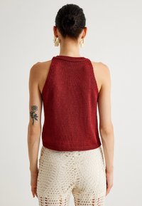 Bakvy av en ärmlös, stickad burgundy topp med ribbad halsringning och kant, tillsammans med virkade vita byxor som visar utskurna mönster.