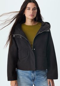 Čierna kapucňová bunda so zipsom vpredu, dvoma prednými vreckami a mierne skráteným strihom, nosená cez olivovo zelený top na svetlom pozadí.