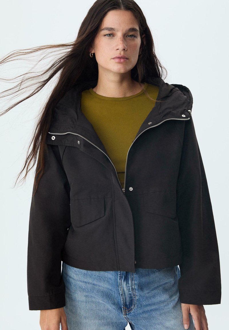 Čierna kapucňová bunda so zipsom vpredu, dvoma prednými vreckami a mierne skráteným strihom, nosená cez olivovo zelený top na svetlom pozadí.