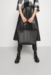 Borsa tote nera con logo in rilievo, forma rettangolare, texture liscia e due manici; indossata con una gonna di pelle e un top a fantasia.