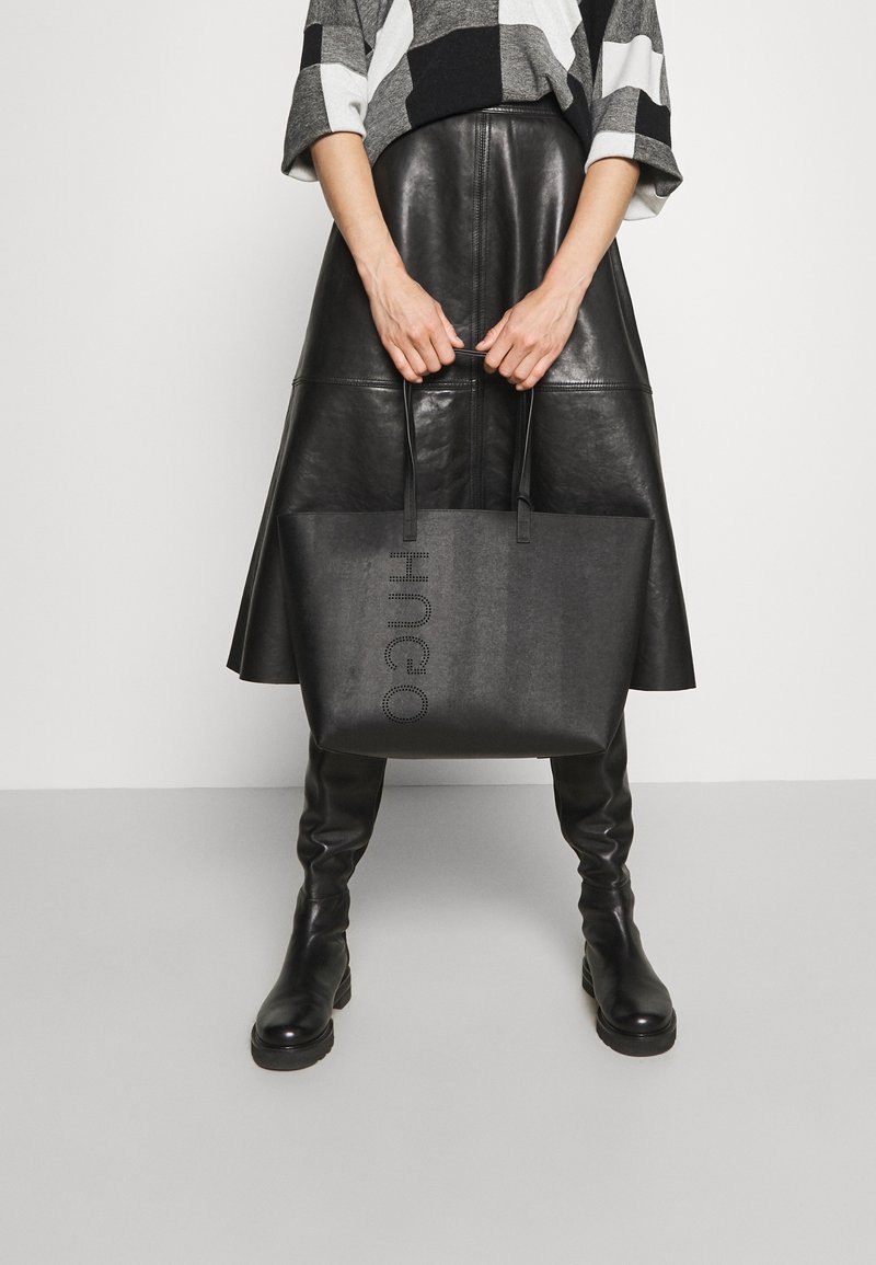 Černá taška typu tote s vyraženým logem, obdélníkového tvaru, hladké textury a dvěma uchy; předváděná s koženou sukní a vzorovaným topem.