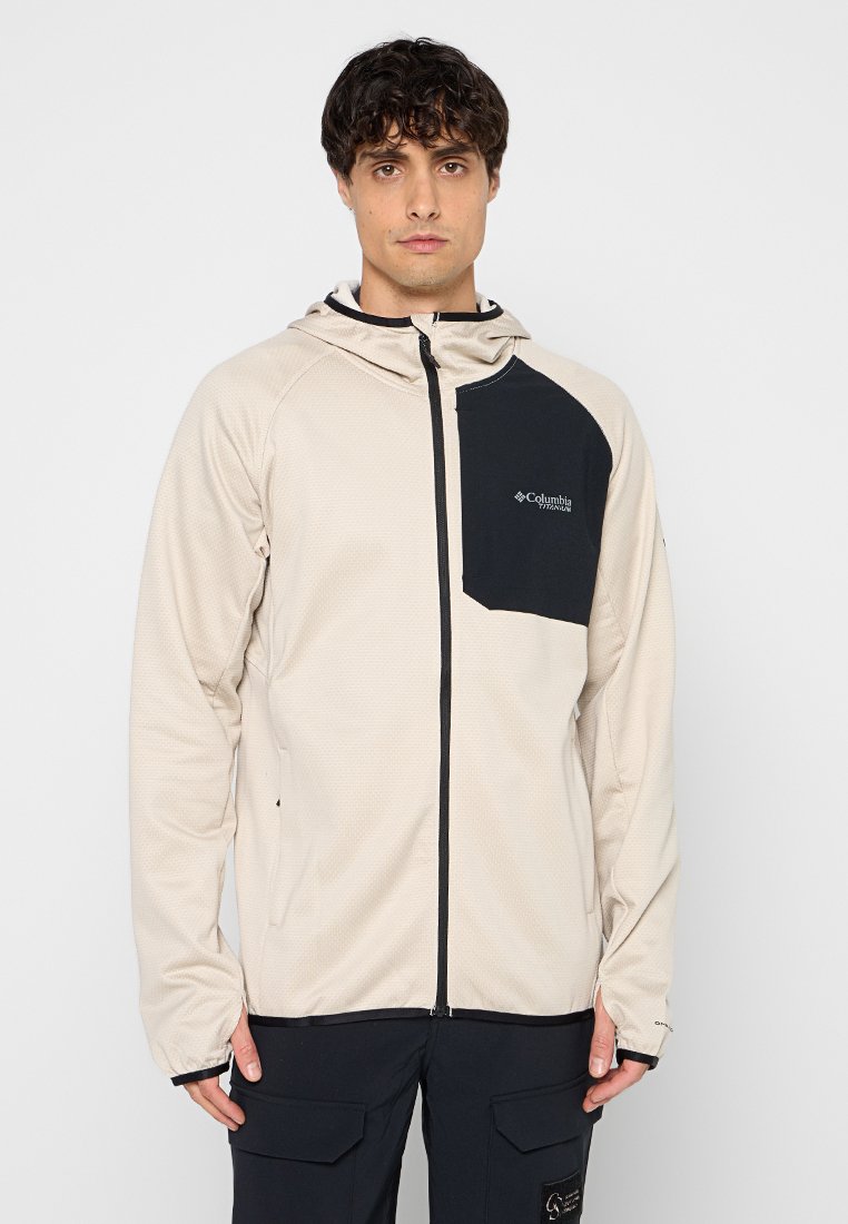Beżowa kurtka z kapturem o teksturowanym wykończeniu, z czarną kieszenią na piersi i kontrastowym zamkiem, zaprojektowana z myślą o aktywnościach na świeżym powietrzu.