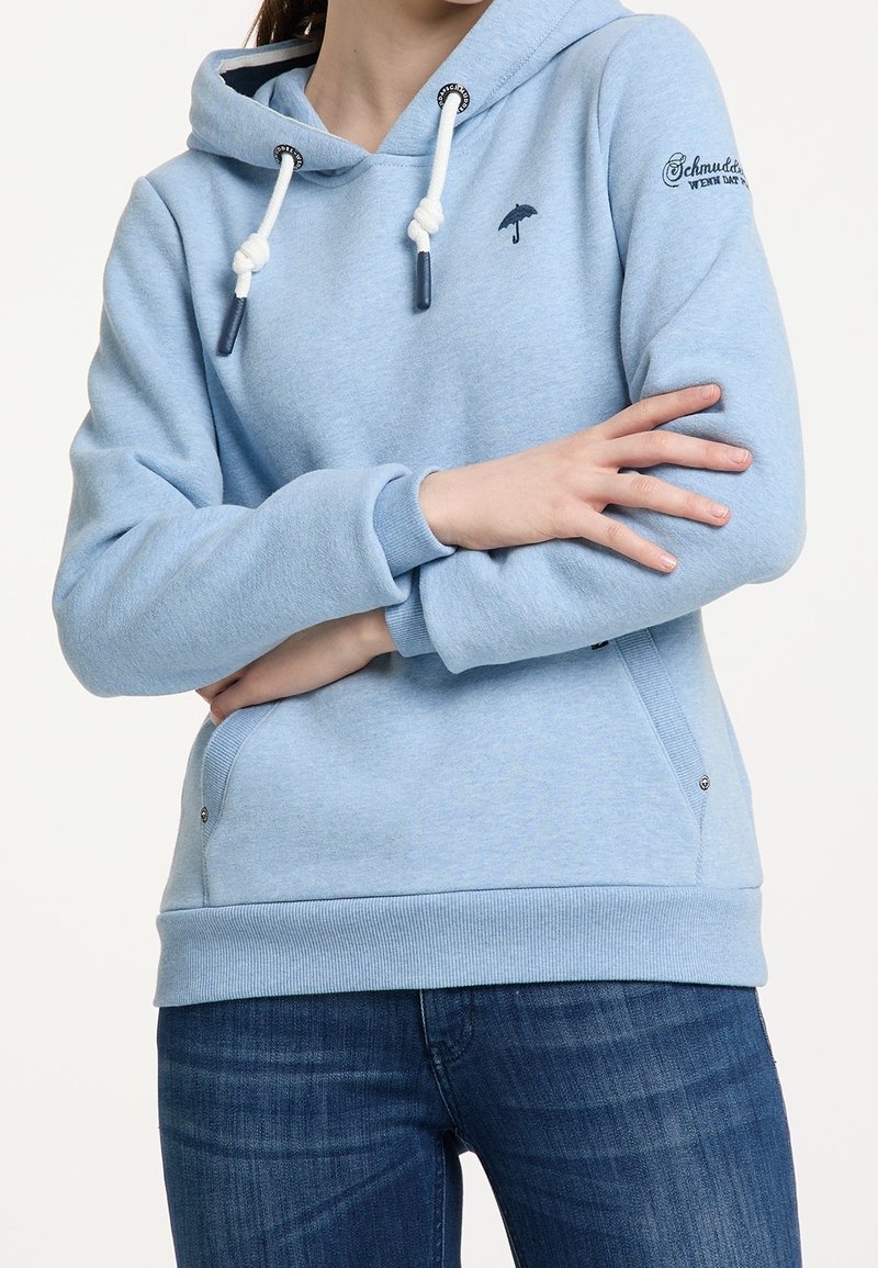 Personne portant un sweat à capuche bleu clair avec des cordons blancs, petit logo parapluie sur la poitrine, texte manuscrit sur la manche et jean bleu, bras croisés.
