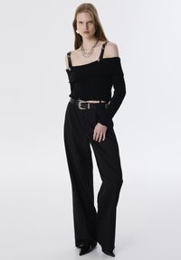 Zwarte off-shoulder trui met ribbeltextuur, gecombineerd met op maat gemaakte zwarte broeken. Opvallende zilveren hardware accenten op de banden en de riem.