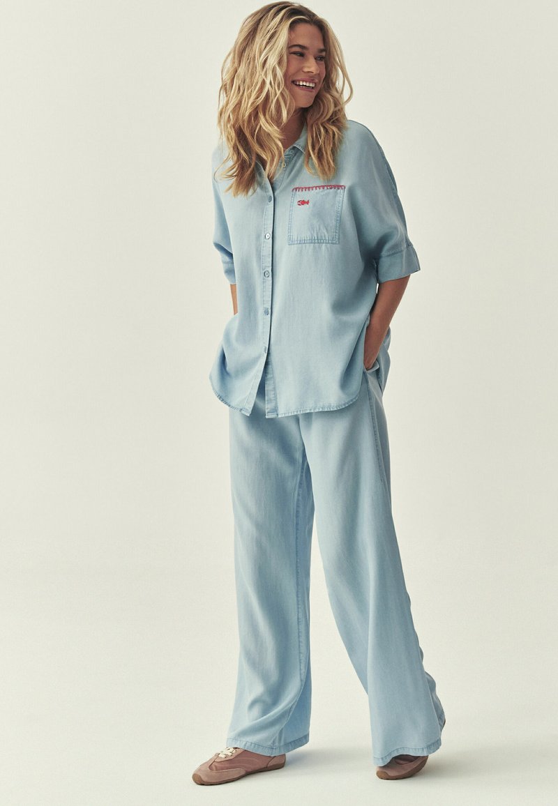 Femme debout, les mains dans les poches, portant une chemise en jean bleu clair ample et un pantalon large avec des baskets marron, souriante et regardant sur le côté.
