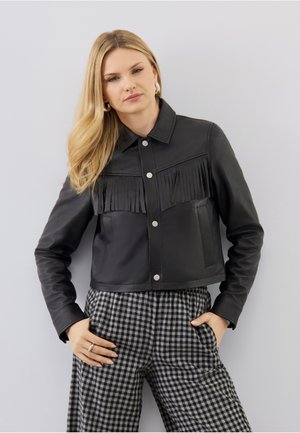 Frau trägt schwarze Lederjacke mit Fransen-Detail und schwarz-weiß karierte Hose, steht vor hellem grauem Hintergrund.
