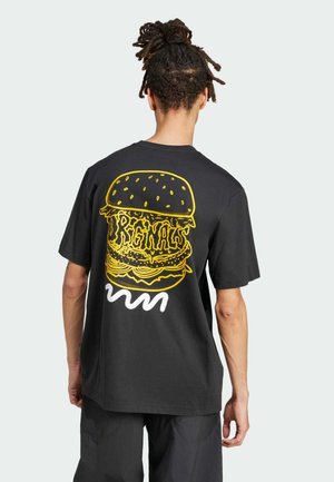 Person med opsat hår iført en sort t-shirt, der viser en stor gul burgergrafik med teksten "REGNALS" på ryggen.