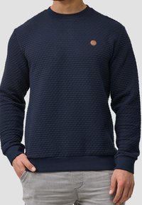 Marineblauwe trui met gestructureerde, patroonstof, geribbelde manchetten en zoom. Voorzien van een klein bruin leren logo op de borst.