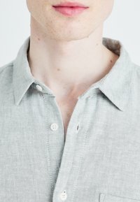 Světle šedá lněná košile s límečkem, s knoflíkovým zapínáním, jemnou texturou a jednou náprsní kapsou.