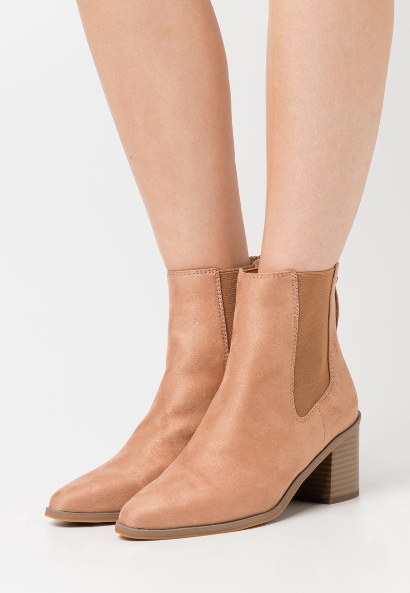 Bottes cheville en daim camel avec bout pointu, panneaux latéraux élastiques et talon empilé. Texture lisse et design minimaliste.