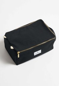Bolsa de viaje de lona negra con cierre dorado, etiqueta blanca con logo y acentos en cuerda color crema. Forma rectangular con base plana.