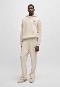 Beige katoenen hoodie met een voorkruiszak en logo, gecombineerd met bijpassende tapered joggingbroek met zijsplitten. Model draagt casual schoenen.