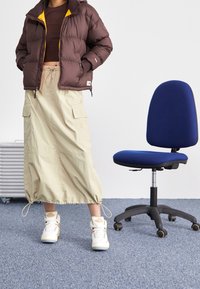 Braune Pufferjacke mit gelbem Futter, beige Cargo-Rock, weiße Sneakers mit Akzenten, steht neben einem blauen Bürostuhl auf grauem Teppich.
