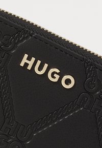 Billetera de cuero negro texturizado con patrones de cadenas en relieve, un logo dorado "HUGO" y un cierre con cremallera en la parte superior.