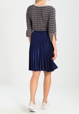 Vrouw die een zigzagpatroon 3/4 mouwen top draagt en een geplooide metallic marineblauwe rok tot op de knie met beige sneakers, staand met de rug naar voren.