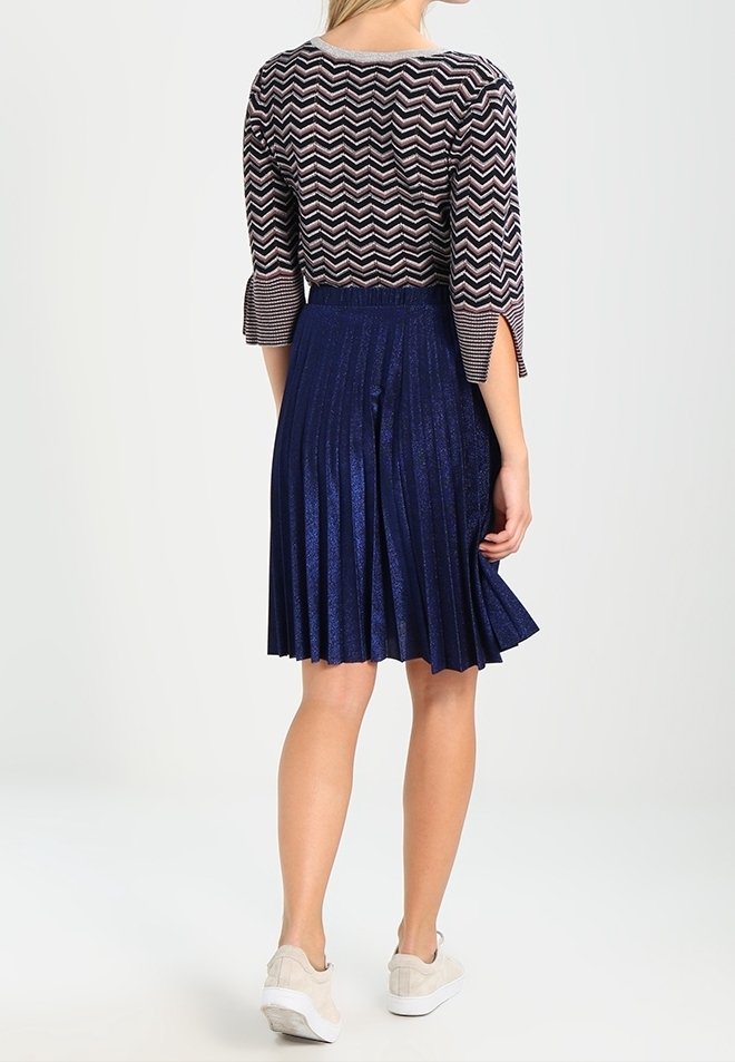 Femme portant un haut manches 3/4 à motif zigzag et une jupe plissée métallique bleu marine longueur genou avec des baskets beiges, debout, dos tourné.
