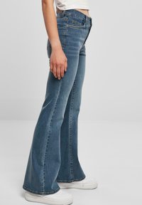 Flared blue denim jeans with a high waist, featuring a faded wash, visible stitching, and five-pocket design. Paired with white sneakers.