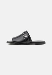 Svart skidsandal med en glänsande finish, som har ett brett band och en silverfärgad logotypaccent. Sulan är platt och tillverkad av hållbart material.