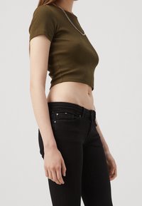 Top corto de canalé verde olivo con mangas cortas, combinado con jeans ajustados negros. Acentos de botón metálico y cremallera en los jeans.