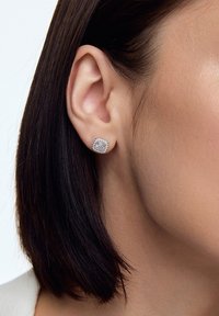 Kwadratowe kolczyki-wkrętki z przezroczystym, błyszczącym kamieniem otoczonym metalową oprawą w srebrnym odcieniu, noszone na uchu.