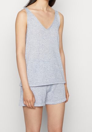 Mujer con top sin mangas gris claro de cuello en V y pantalones cortos a juego, de pie frente a un fondo blanco liso.