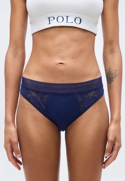Femme portant un slip bleu marine à bordure en dentelle et un crop top blanc avec le texte "POLO", montrant le ventre et un tatouage sur l'avant-bras gauche.