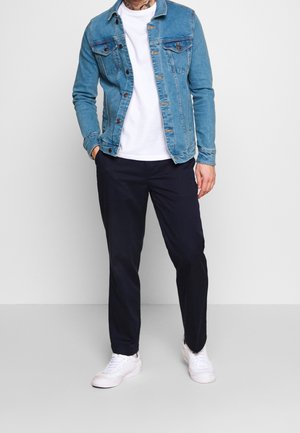 Hombre con chaqueta de mezclilla azul sobre camiseta blanca, pantalones negros y zapatillas blancas, de pie contra un fondo claro y liso.