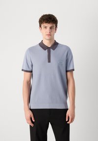 Polo azul de manga curta com um padrão texturizado, gola escura em contraste e um logotipo circular no lado esquerdo do peito, combinado com calças pretas.