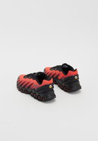 Baskets de sport rouges et noires avec un dessus en maille, un design de vague distinctif et une semelle large en coussin d'air arrondi. Présente un logo à l'arrière.