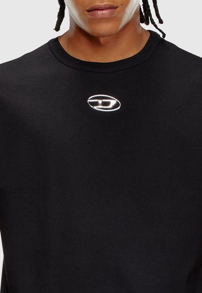 T-shirt noir avec un col rond, arborant un logo ovale en argent sur la poitrine. Le tissu semble lisse et doux au toucher.