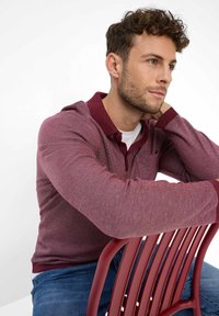 Polo shirt bordeaux a maniche lunghe con tessuto testurizzato, colletto a contrasto e tasca. Indossato con jeans blu, seduto su una sedia rossa.