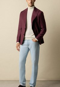 Burgundowa marynarka w prążki, beżowy golf, jasnoniebieskie dżinsy i ciemne buty. Marynarka ma wyłogowy kołnierz i dwa guziki.