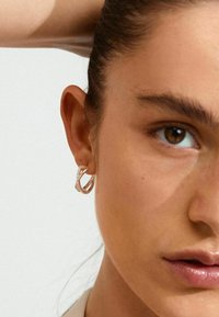 Pendiente grande en forma de aro retorcido en metal tono plata con un acabado texturizado. Primer plano del rostro de un modelo que muestra la oreja y el detalle del pendiente.