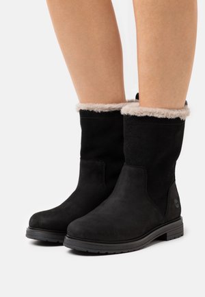 Botines de ante negro con un suave puño de piel de pelo beige y una suela de goma texturizada. Presentan un logotipo en el costado y un acabado liso.