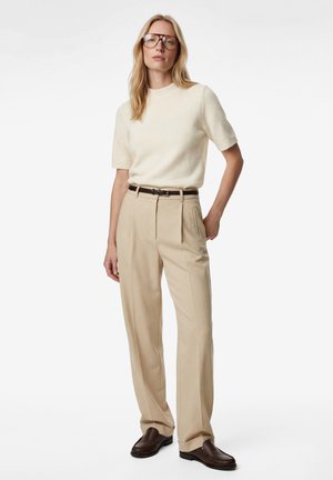 Femme portant un pantalon beige taille haute, un pull crème à manches courtes, une ceinture marron, des chaussures marron et de grandes lunettes rondes, debout avec une main dans la poche.
