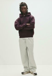 Felpa con cappuccio bordeaux, vestibilità ampia. Pantaloni della tuta grigi chiari, silhouette morbida. Scarpe da ginnastica nere e grigie con dettagli in rete. Sfondo minimale.