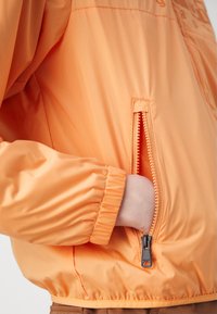 Pomarańczowa kurtka nylonowa z zamkiem błyskawicznym w kieszeni, elastycznymi mankietami i gładką teksturą. Projekt jest prosty z minimalnymi akcentami.