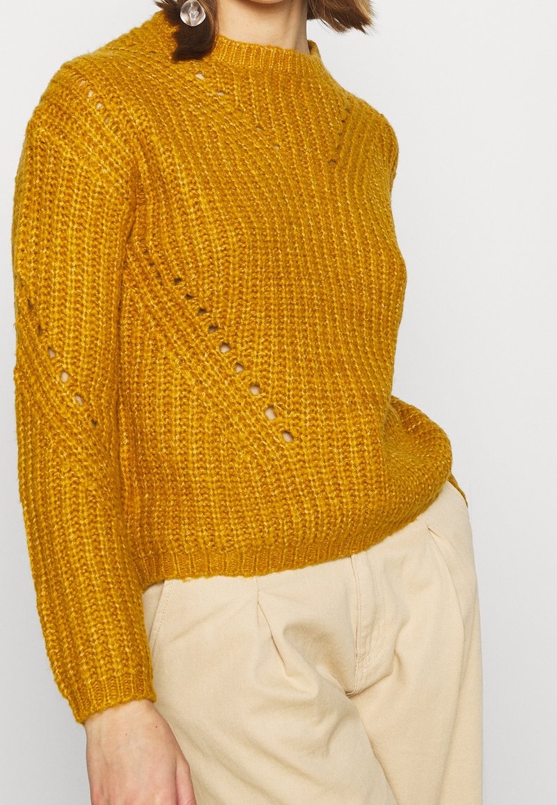 Femme portant un pull en maille épaisse jaune moutarde avec des détails en œillets diagonaux, associé à un pantalon taille haute beige.