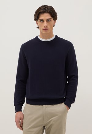 Giovane uomo con capelli castani che indossa un maglione girocollo blu navy sopra una camicia bianca e pantaloni beige, in piedi contro uno sfondo semplice.